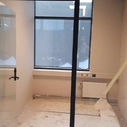 DUDA FINISH - Przygotowanie do malowania biura: podłoga zabezpieczona folią malarską, widoczny grzejnik i okno w czarnej ramie, sufit podwieszany z widocznymi elementami konstrukcyjnymi.