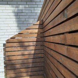 DUDA FINISH - Kąt ujęcia na drewniany płot z desek o brązowym odcieniu, przylegający do białej ściany z cegły, z widocznym cieniem rzucanym przez słońce.