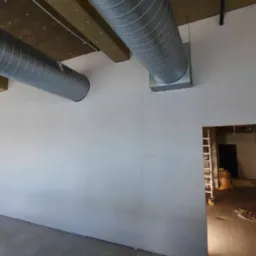 Wnętrze w trakcie montażu płyt kartonowo-gipsowych na ścianach, widoczne metalowe rury wentylacyjne pod sufitem i otwór drzwiowy z drabiną w tle.