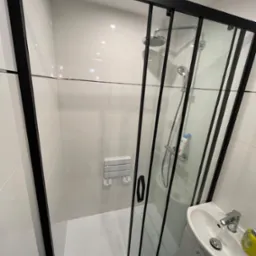 Nowoczesna łazienka z kabiną prysznicową z czarnymi ramami, białe płytki na ścianach, umywalka z szafką i składane siedzisko prysznicowe.