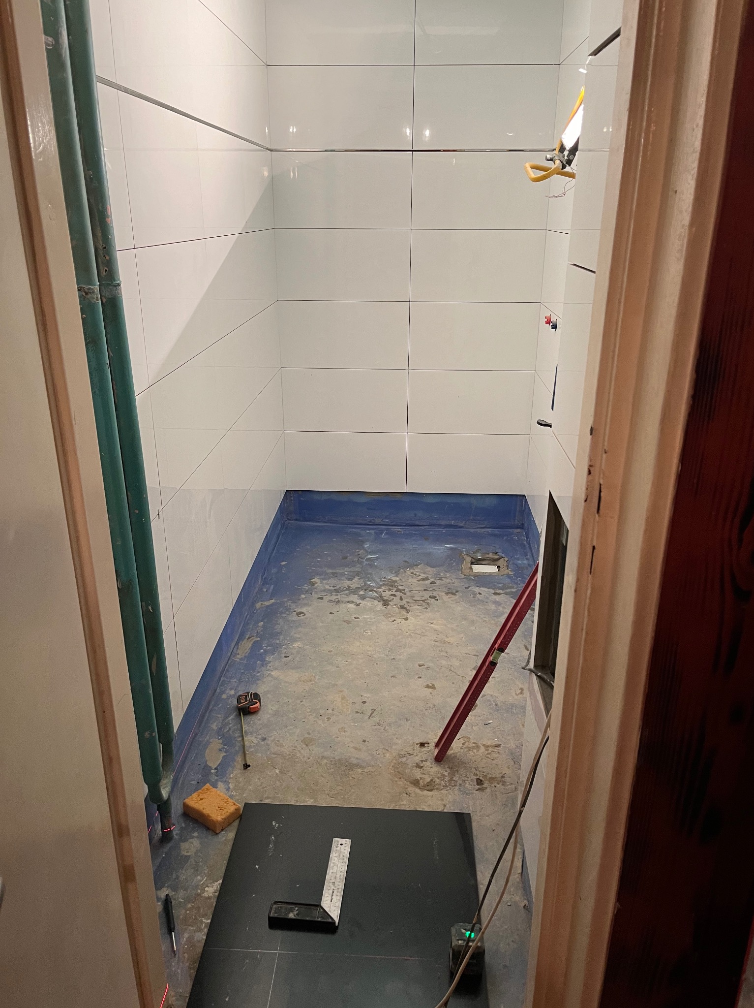 Łazienka w trakcie przygotowania do układania płytek: widoczne białe płytki na ścianach, niebieska hydroizolacja na podłodze, poziomica, kątownik, gąbka, rury i laser krzyżowy.