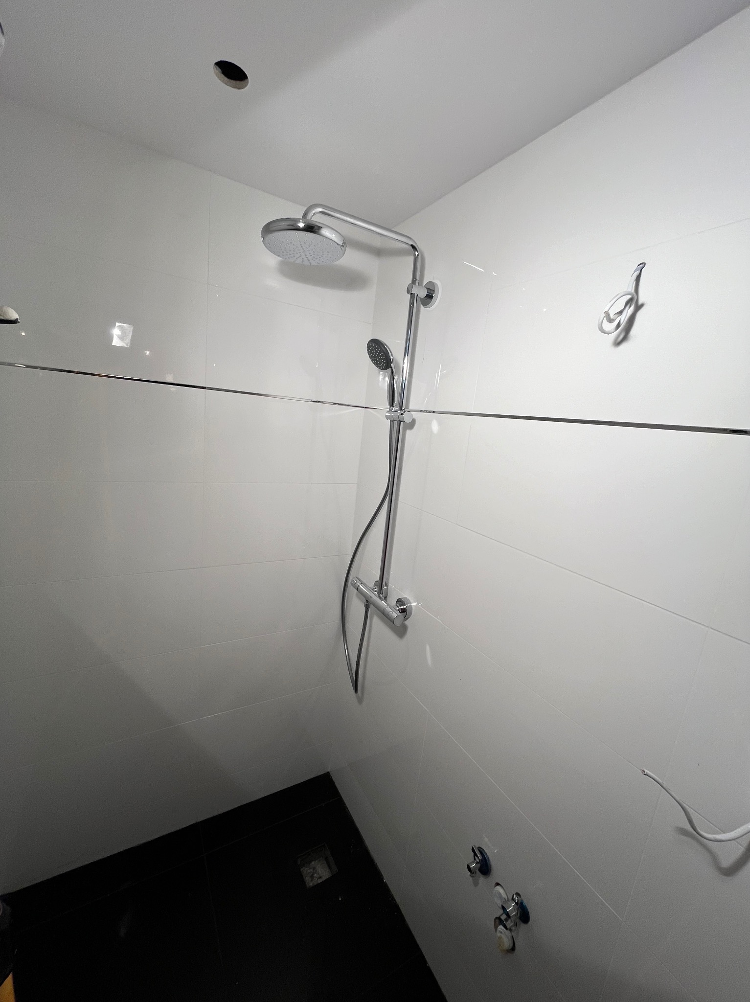 Nowoczesna kabina prysznicowa z chromowaną armaturą, widoczne białe płytki ścienne z cienką listwą dekoracyjną i czarne płytki podłogowe, niedokończone podłączenie elektryczne w rogu.