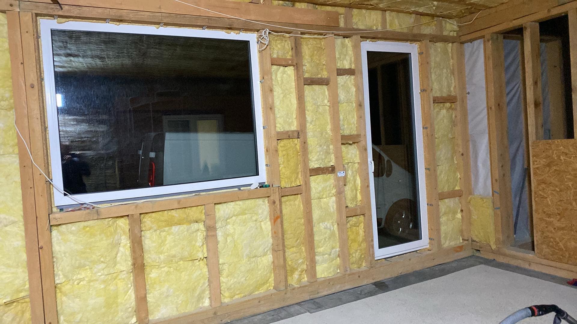 Ściana w trakcie ocieplania wełną mineralną, widoczne drewniane ramy konstrukcyjne, okno z białymi ramami oraz drzwi balkonowe, w tle zaparkowany samochód.