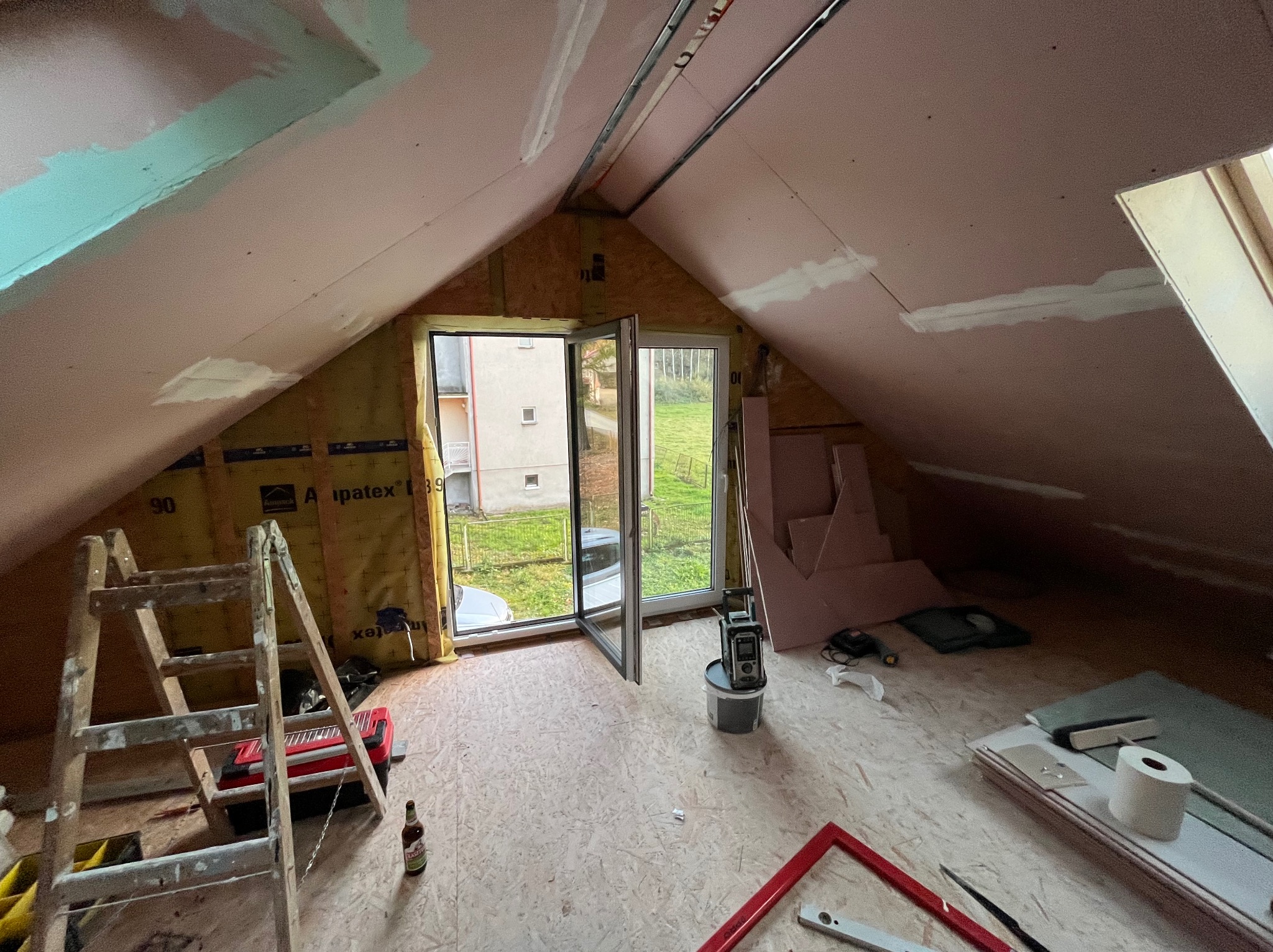 Adaptacja poddasza: widok na ocieplone ściany skośne z oknami, częściowo pokryte płytami kartonowo-gipsowymi, drabina, narzędzia i materiały budowlane rozłożone na podłodze z płyt OSB.