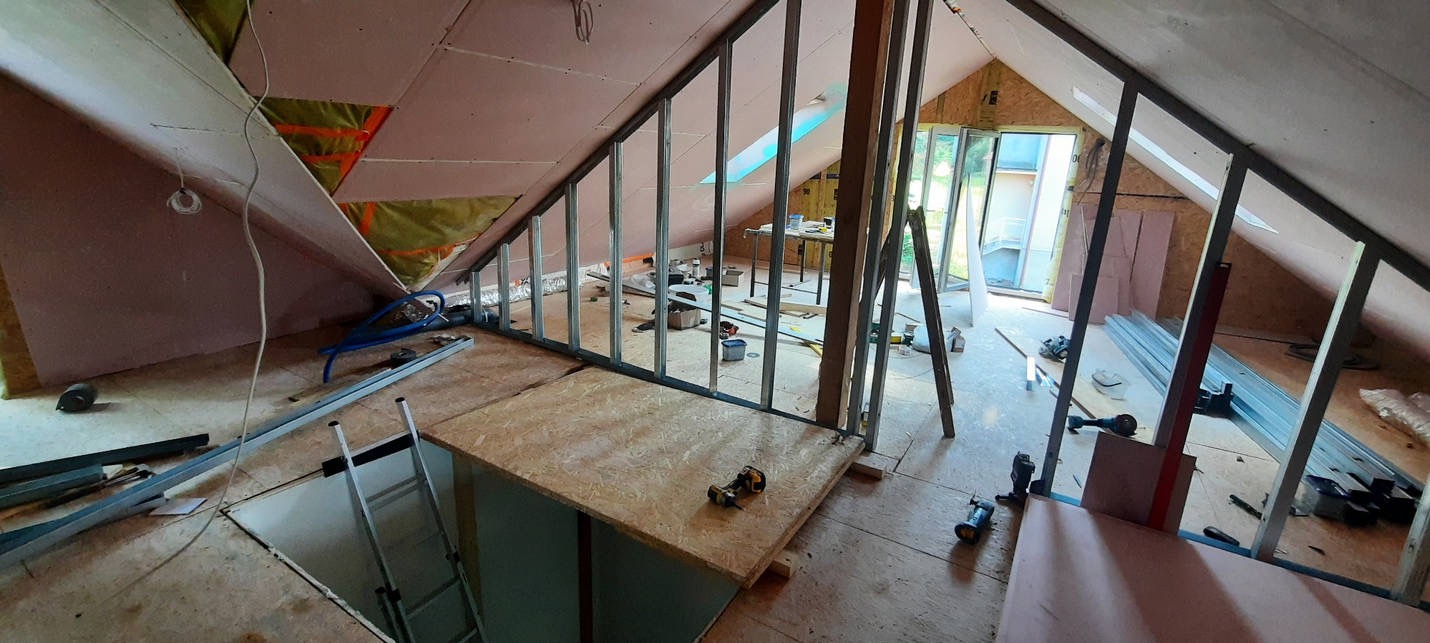 Adaptacja poddasza: widok na szkielet z profili stalowych, częściowo obłożony płytą karton-gips, z widoczną izolacją z wełny mineralnej i otworem w podłodze z drabinką.