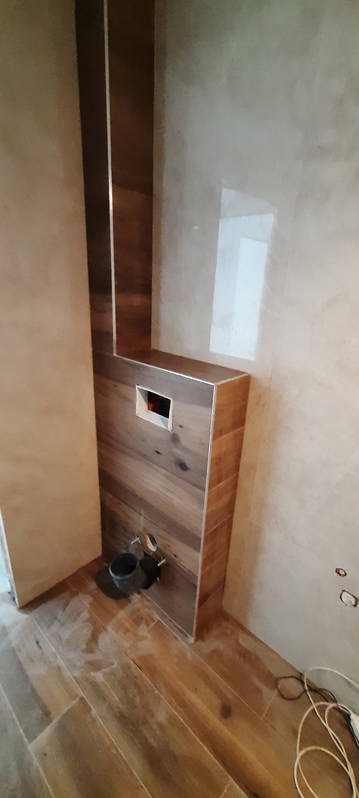 Stelaż podtynkowy WC obudowany płytkami imitującymi drewno, z widocznymi przyłączami i częściowo wykończoną ścianą w beżowym odcieniu, na podłodze ułożone płytki podłogowe o strukturze drewna.