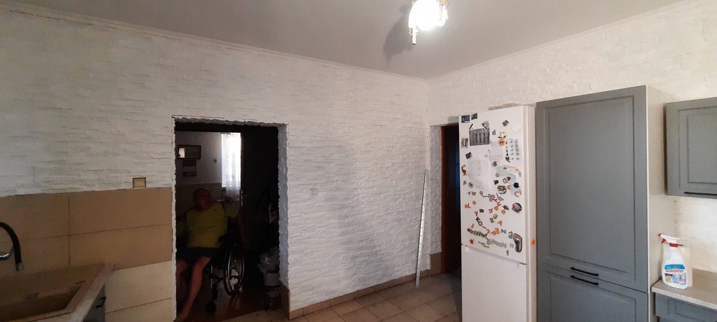 Wykończona kuchnia z białymi ścianami imitującymi cegłę, szare szafki kuchenne, biała lodówka z magnesami i osobą na wózku inwalidzkim widoczną w przejściu do innego pomieszczenia.