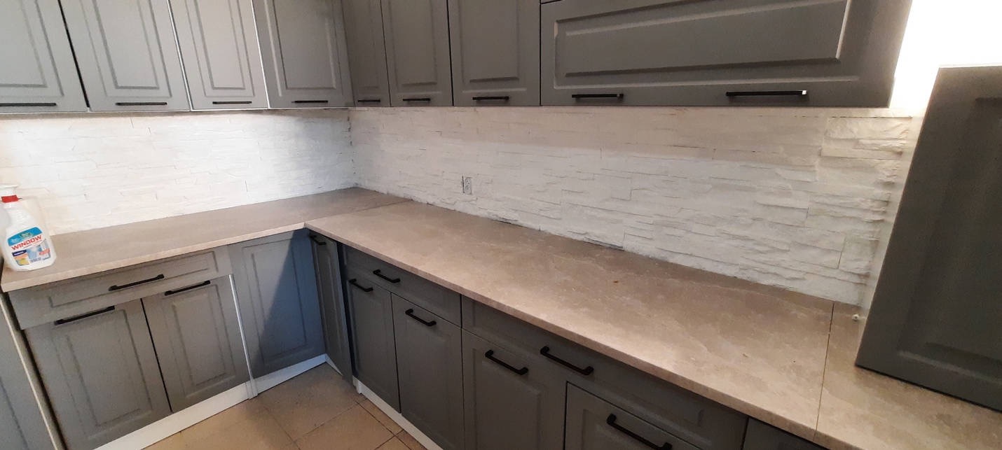 Szare szafki kuchenne z czarnymi uchwytami, beżowy blat i ściana wyłożona dekoracyjną białą cegłą, widoczny środek do czyszczenia okien