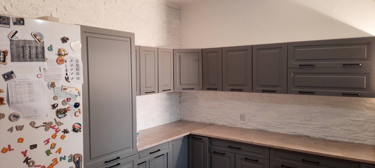 Szare szafki kuchenne z czarnymi uchwytami, biała ściana z imitacją kamienia, lodówka z magnesami w tle.