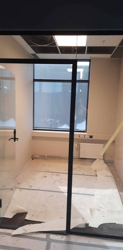 Przygotowanie do malowania biura: podłoga zabezpieczona folią malarską, widoczny grzejnik i okno w czarnej ramie, sufit podwieszany z widocznymi elementami konstrukcyjnymi.