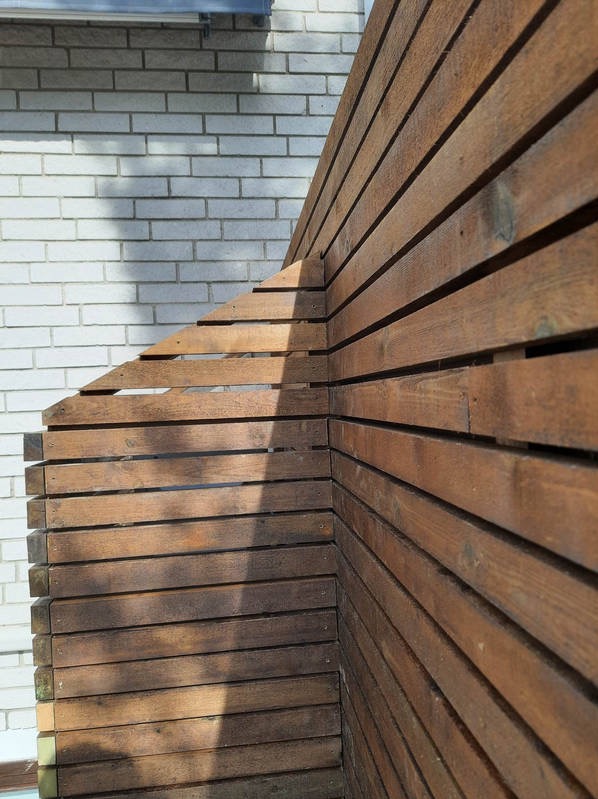 Kąt ujęcia na drewniany płot z desek o brązowym odcieniu, przylegający do białej ściany z cegły, z widocznym cieniem rzucanym przez słońce.