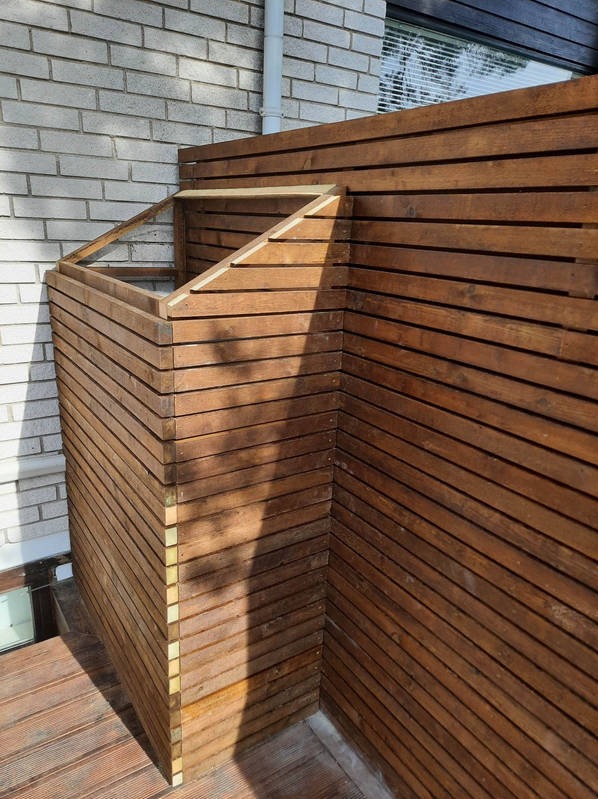 Drewniana zabudowa tarasu z poziomych desek, tworząca osłonę na śmietnik lub schowek, przylegająca do murowanej ściany budynku.