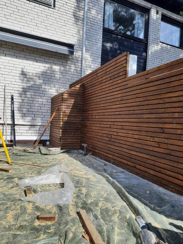 Montaż drewnianego ogrodzenia panelowego przy budynku z jasnej cegły, widoczna poziomica i wkrętarka na folii ochronnej.