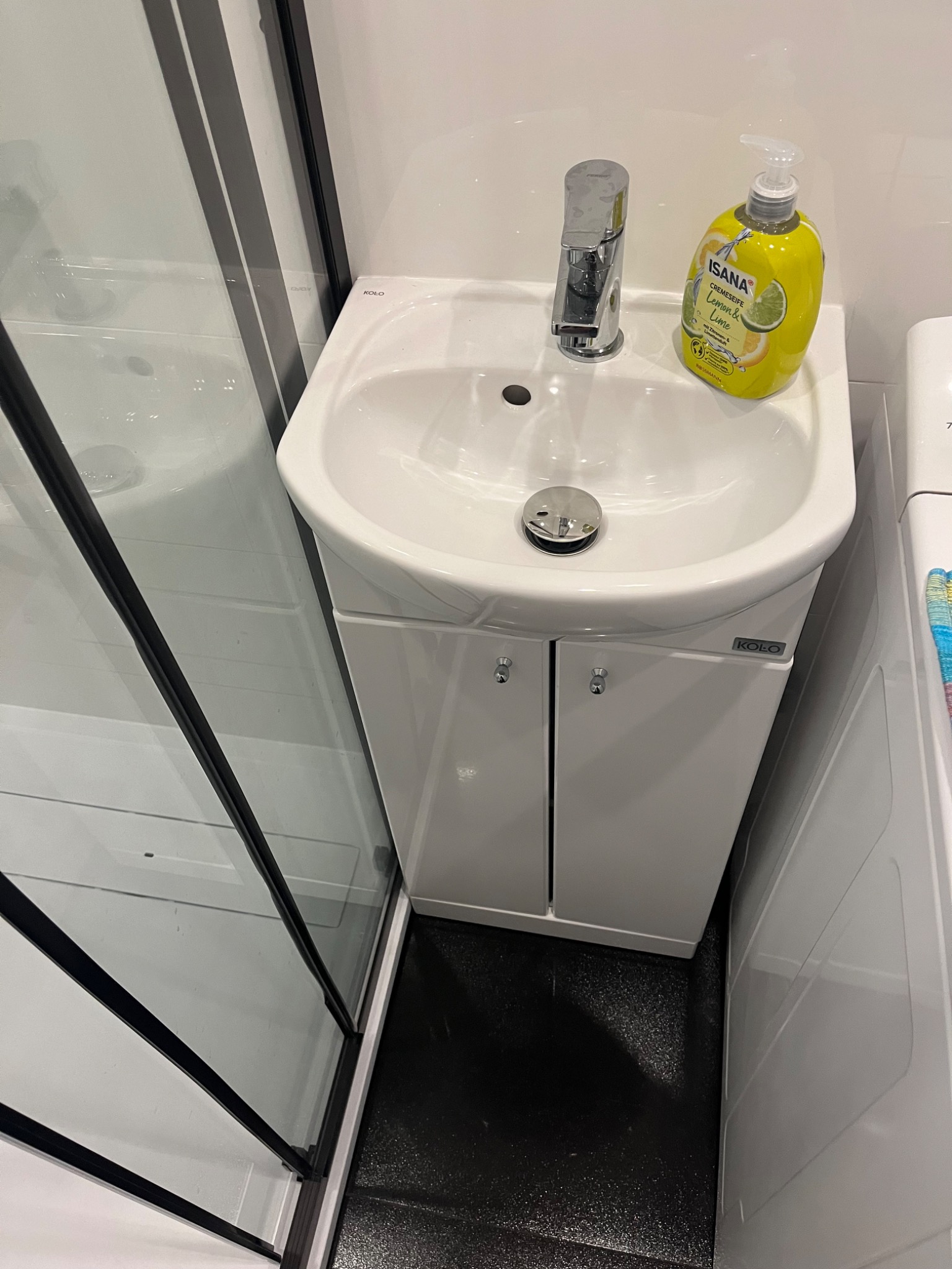 Nowoczesna biała umywalka z szafką marki Koło w łazience, bateria chromowana, obok kabina prysznicowa z czarnymi profilami i butelka mydła Isana Lemon&Lime.