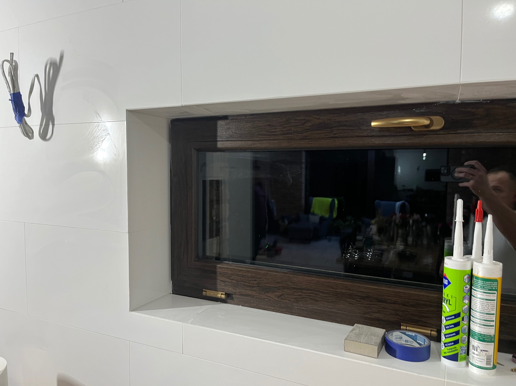 Białe płytki ścienne w łazience z oknem o ciemnobrązowej ramie. Na parapecie okiennym widoczne silikony, taśma malarska i gąbka szlifierska. Na ścianie zwisają przewody elektryczne.