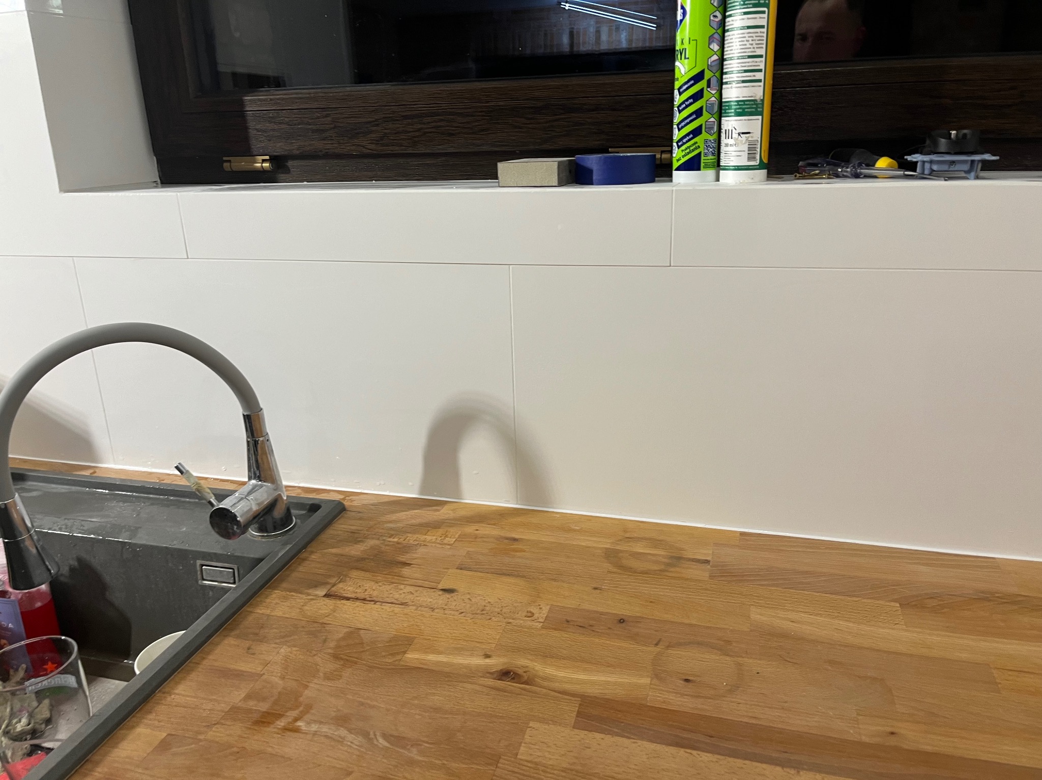 Szary zlewozmywak z baterią na drewnianym blacie pod oknem z białymi płytkami, widoczne ślady wody i akcesoria montażowe.