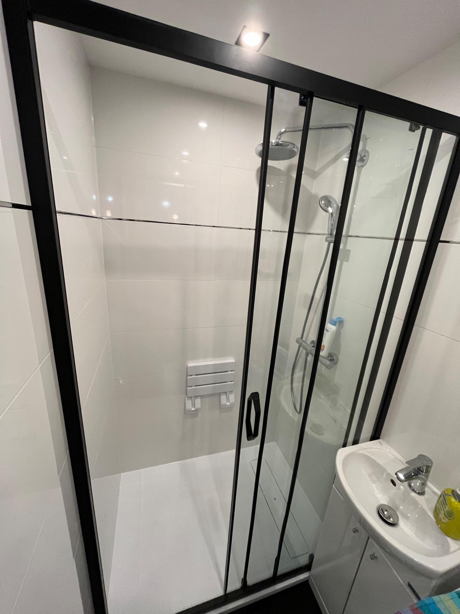 Nowoczesna łazienka z kabiną prysznicową z czarnymi ramami, białe płytki na ścianach, umywalka z szafką i składane siedzisko prysznicowe.