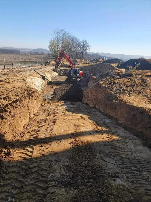 Wykop liniowy w ziemi z widocznymi śladami opon i pracującymi koparkami w tle, wykonany w słoneczny dzień. Widok z góry na dół.