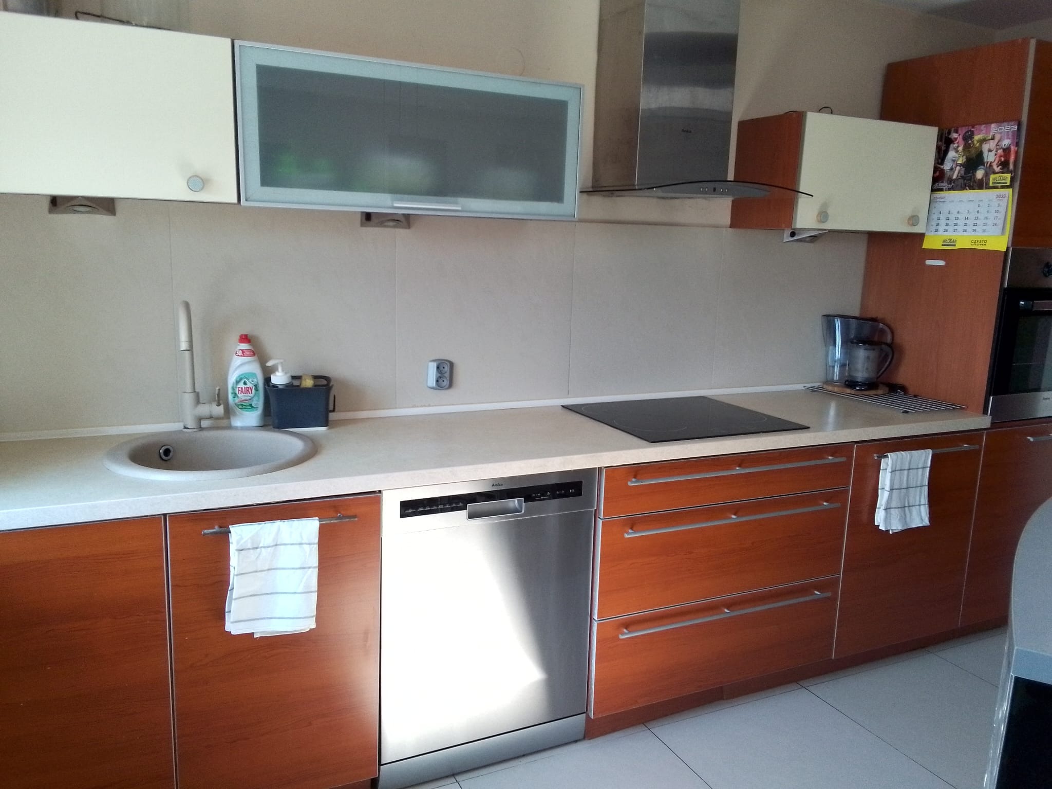 Uporządkowana kuchnia z drewnianymi szafkami, szarą armaturą i sprzętami AGD, z widocznym płynem do naczyń Fairy i kalendarzem na ścianie.