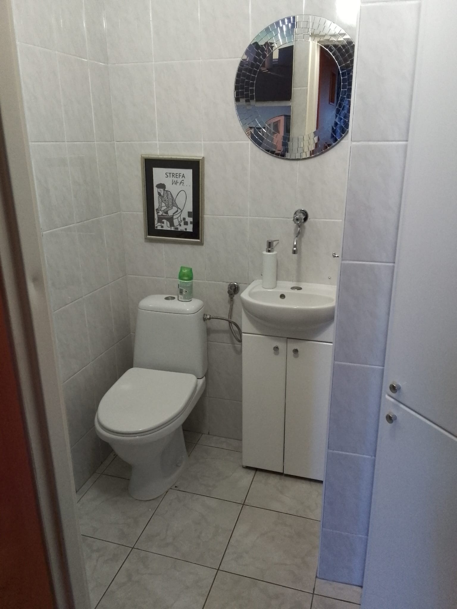 Mała łazienka z białą ceramiką, umywalką z szafką, toaletą z zielonym odświeżaczem powietrza, okrągłym lustrem z mozaiki i obrazkiem z napisem 'Strefa Wi-Fi'.