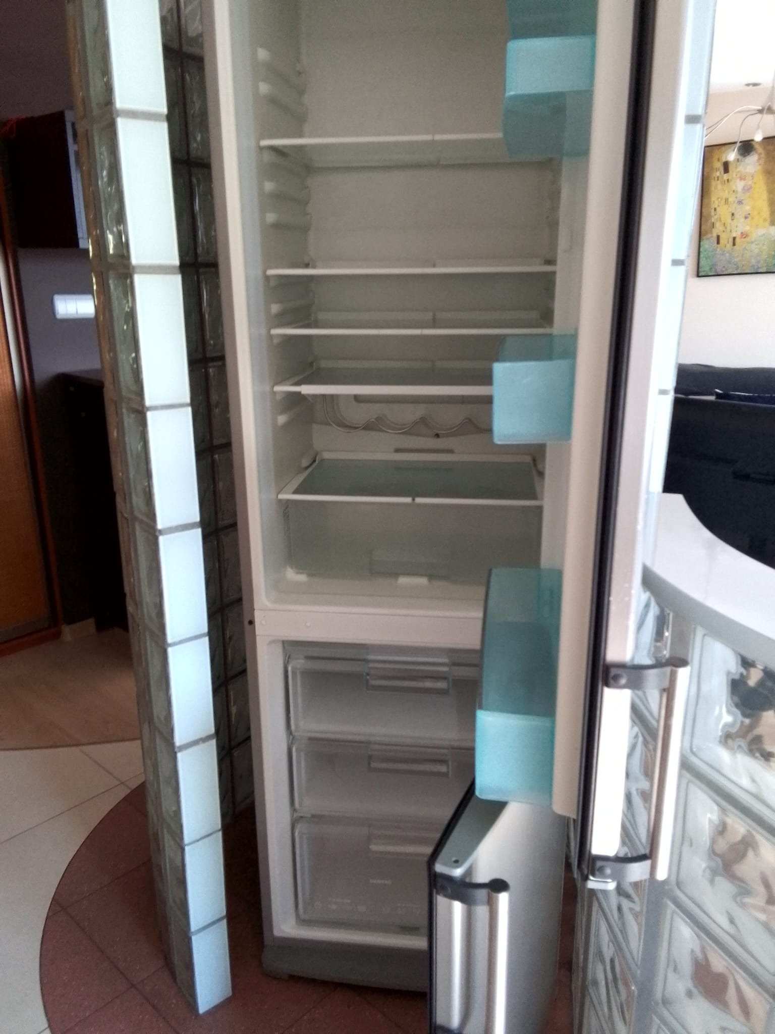 Pusta lodówka z otwartymi drzwiami, widoczne półki i szuflady, obok ściana z luksferów, w tle obraz Gustava Klimta.