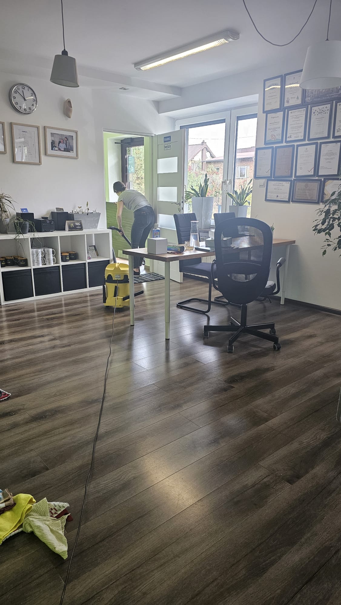 Osoba odkurza podłogę w biurze za pomocą żółtego odkurzacza przemysłowego; widoczne biurko, krzesła, półki z dekoracjami i dyplomy na ścianie.