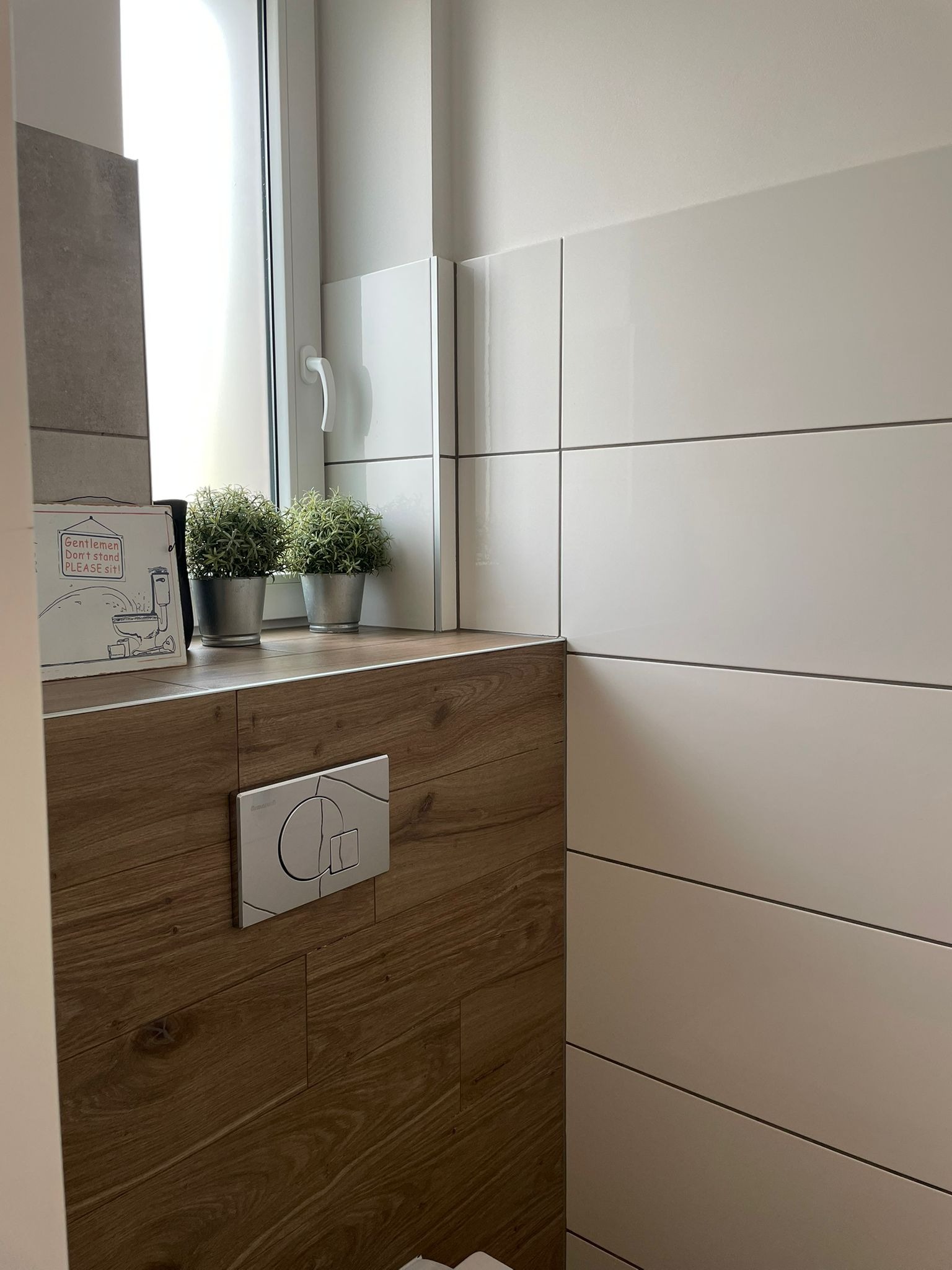 Nowoczesna toaleta z drewnianą zabudową, białymi płytkami, dwoma małymi roślinami w metalowych doniczkach oraz zabawnym rysunkiem z napisem 'Gentlemen Don't stand PLEASE sit!'