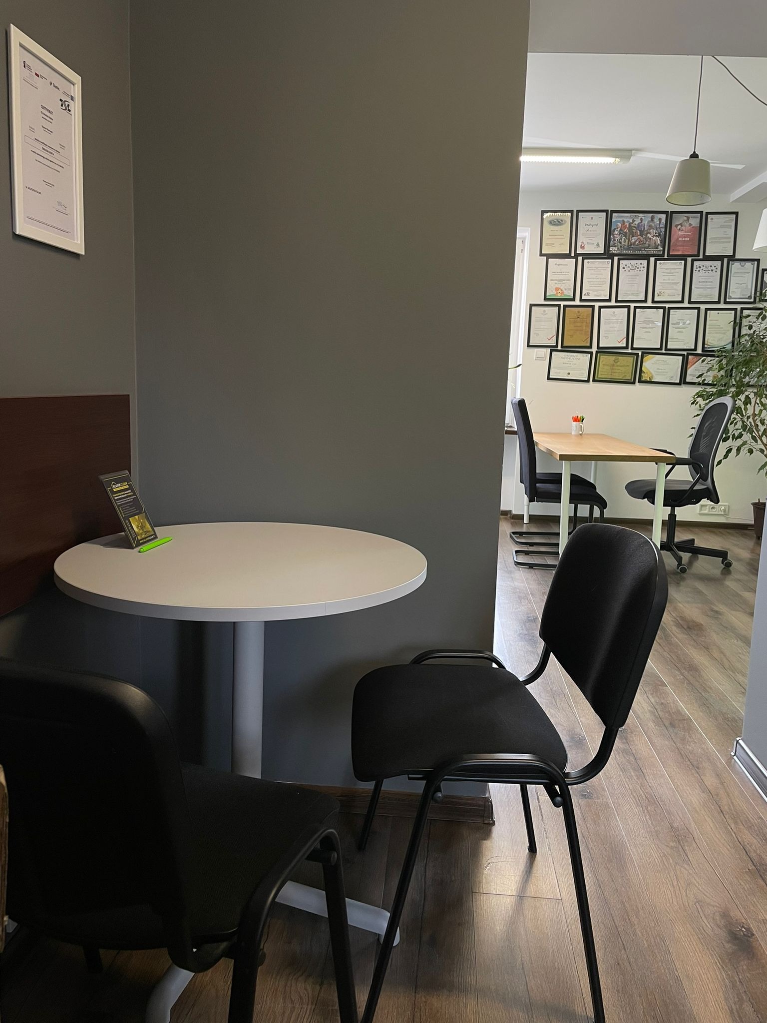 Wnętrze biura z okrągłym stolikiem, czarnymi krzesłami i ścianą z certyfikatami w tle, widoczne drewniane panele podłogowe i broszura informacyjna na stole.