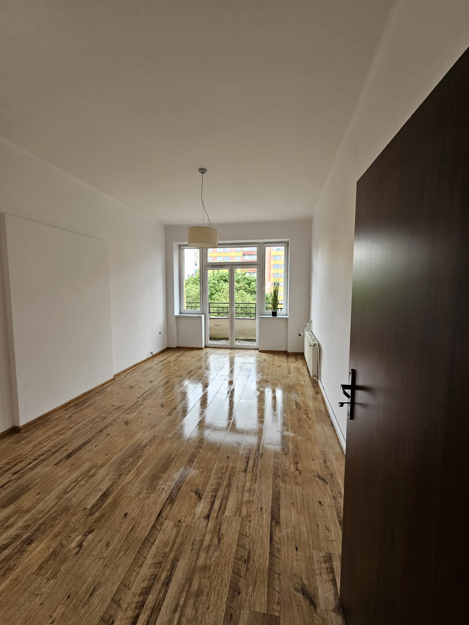 Pusty pokój z drewnianą podłogą po remoncie, świeżo pomalowane ściany na biało, widok na balkon i zieleń za oknem, drzwi wejściowe z prawej strony.