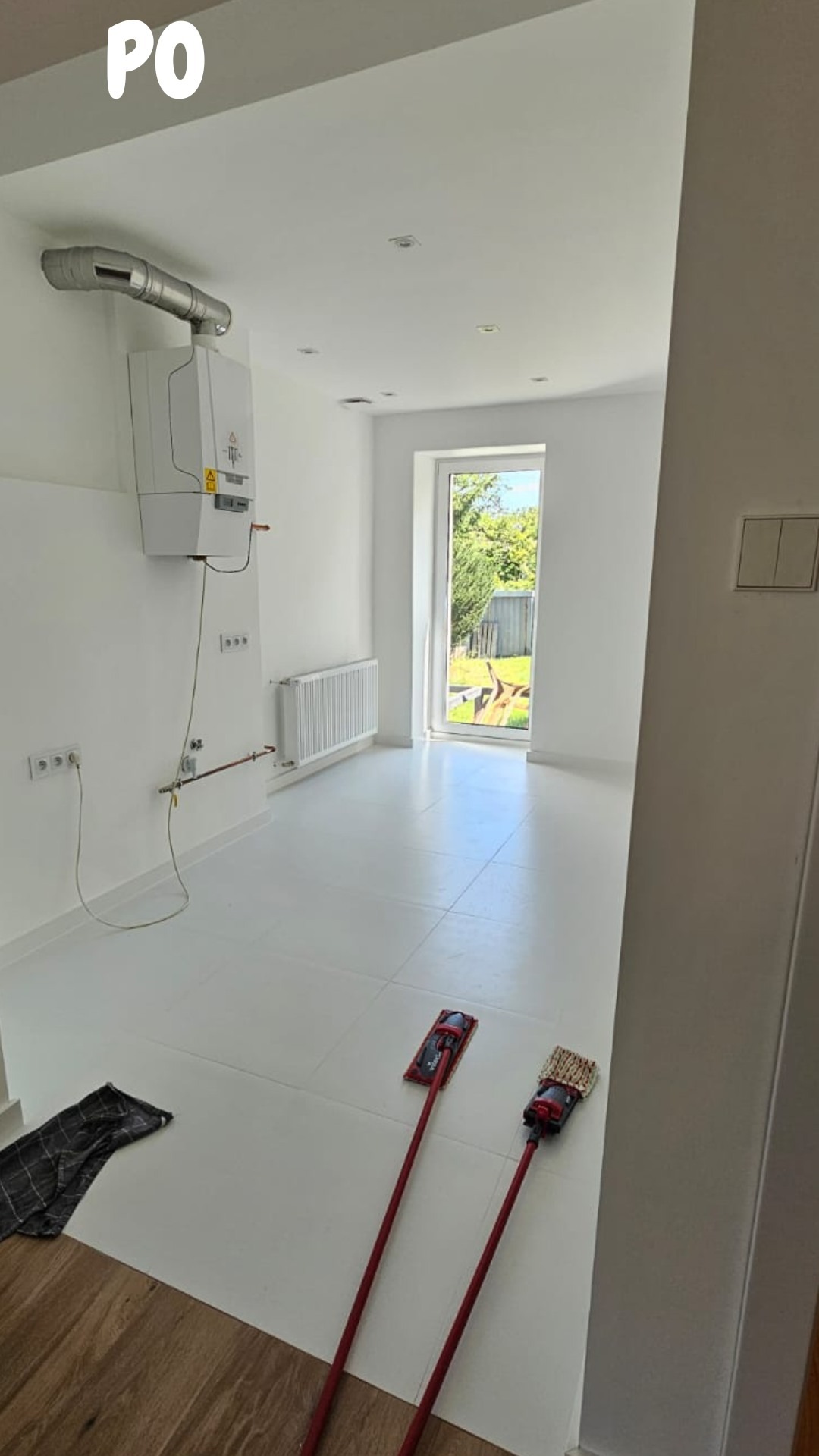 Jasne, przestronne wnętrze po remoncie, z białymi ścianami i podłogą, widoczne dwa mopem płaskie, grzejnik, piec gazowy i okno z widokiem na zieleń.