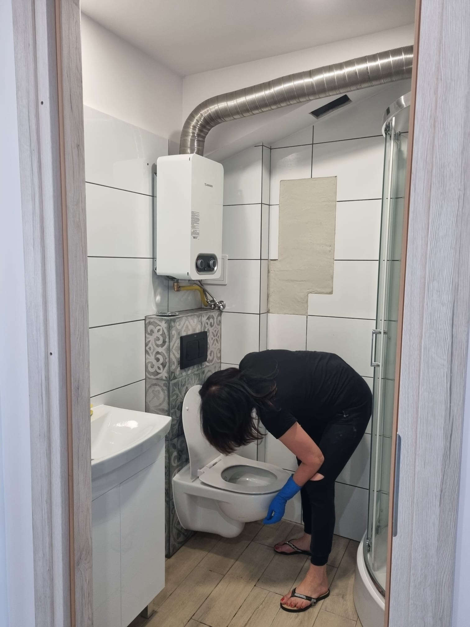 Kobieta w czarnym ubraniu i niebieskich rękawicach czyści toaletę w łazience z białymi płytkami i grzejnikiem wody Junkers.