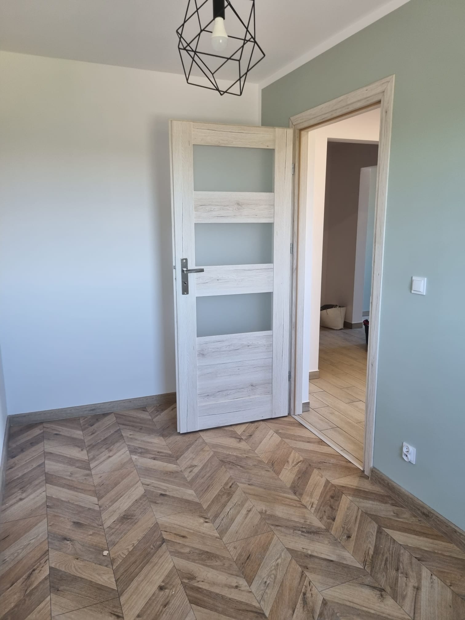 Pomalowane na dwa kolory wnętrze z drewnianą podłogą ułożoną w jodełkę, białe drzwi z czterema szybkami i nowoczesna lampa wisząca.