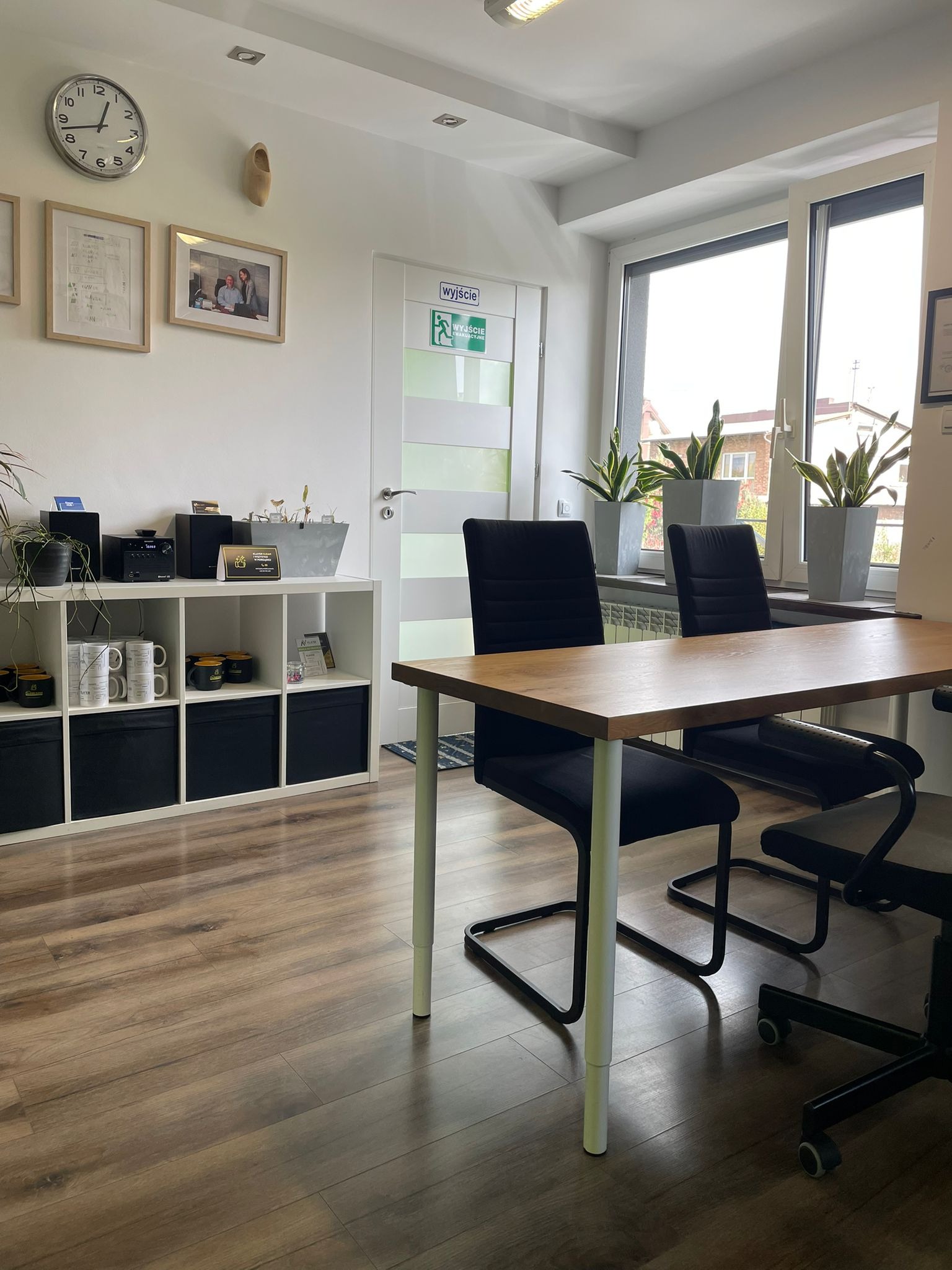 Jasne biuro z drewnianym stołem, czarnymi krzesłami i regałem. Na ścianie zegar, zdjęcia i drewniany but. Okno z roślinami w doniczkach. Podłoga z paneli.