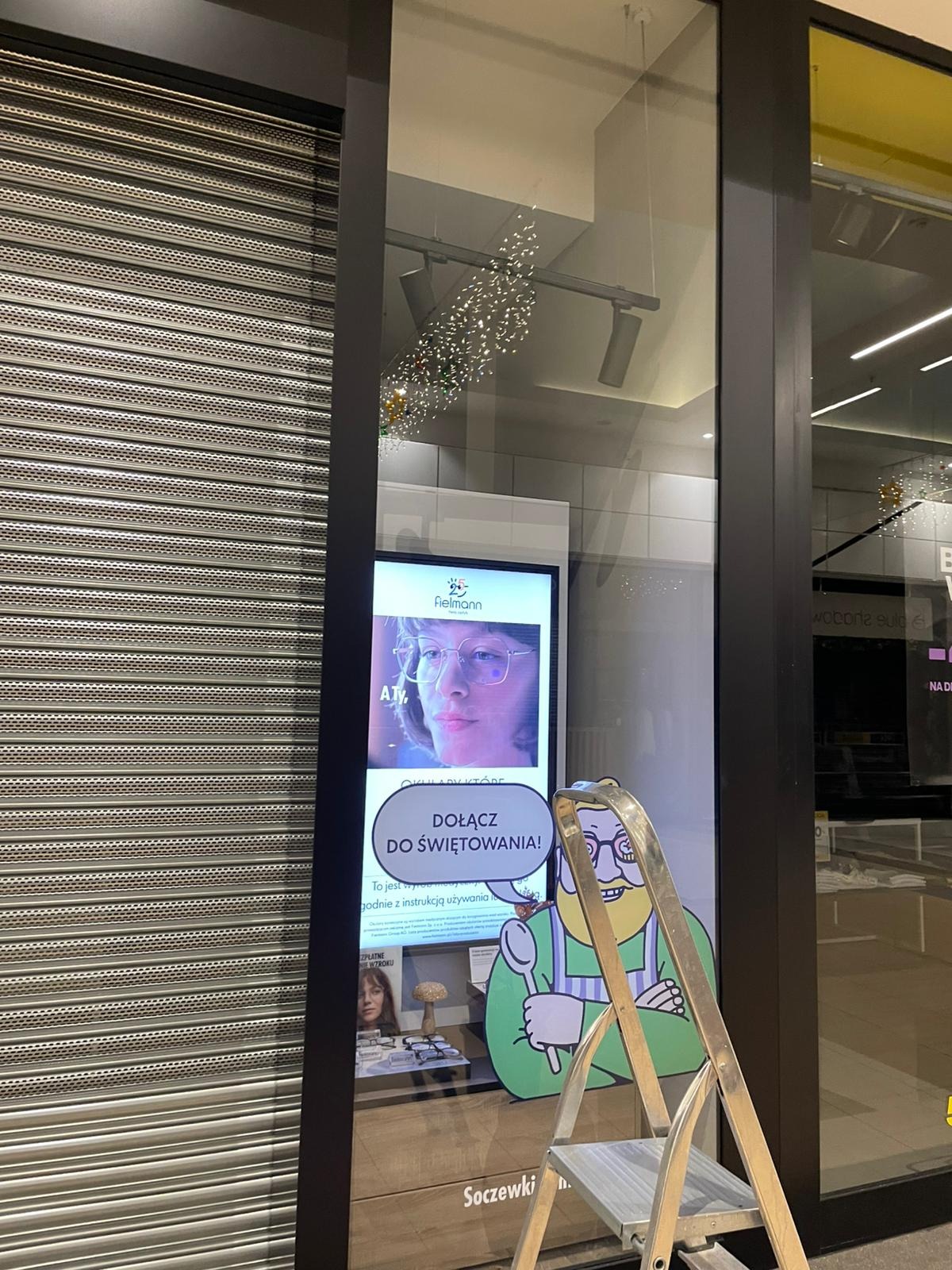 Sklep po godzinach: zamknięta roleta, witryna z reklamą okularów i świąteczną dekoracją, drabina oparta o szybę. Widoczne odbicia światła i otoczenia.