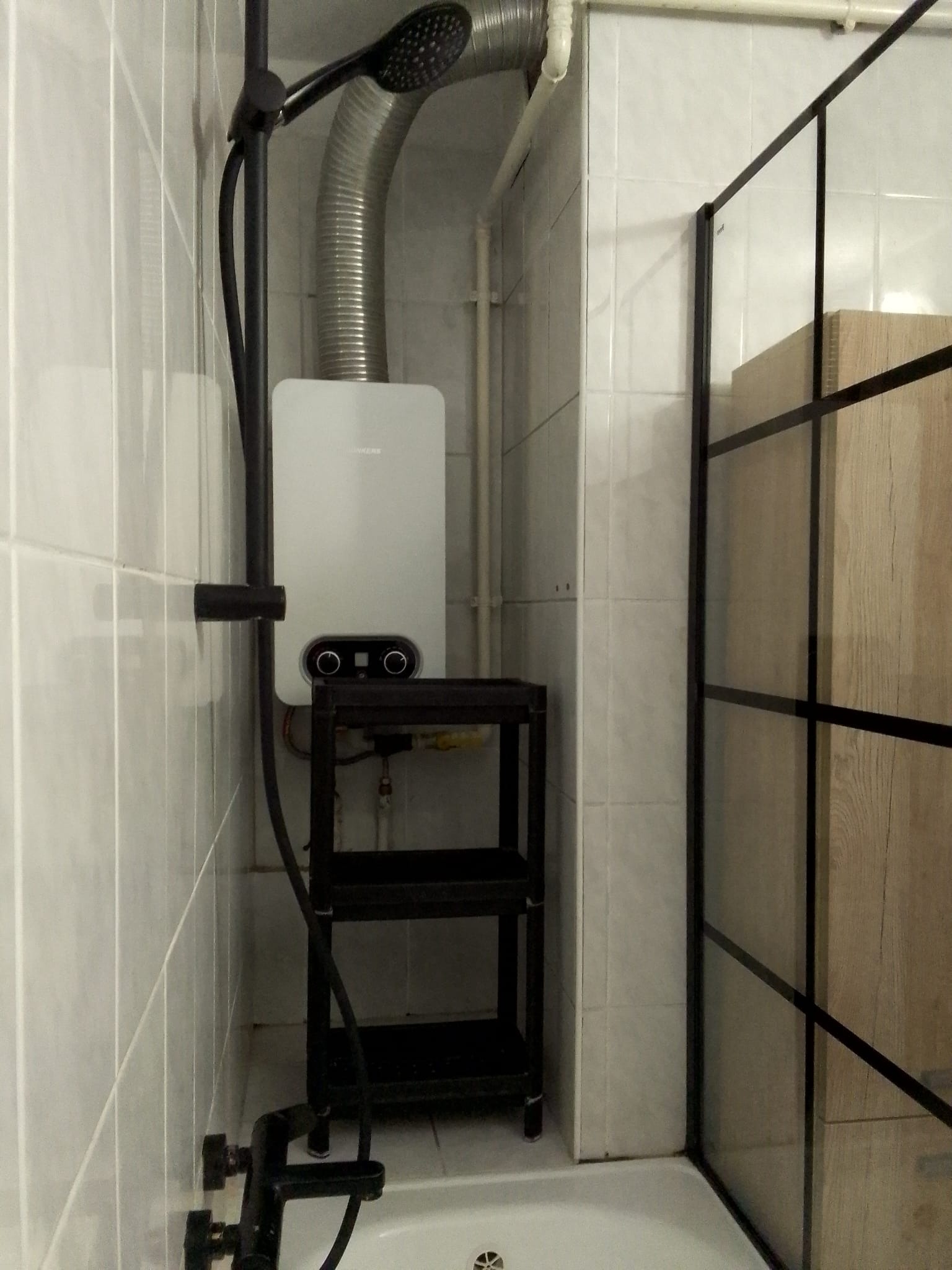 Biały, wiszący piecyk gazowy Junkers z czarnymi pokrętłami, zamontowany w małej łazience z białą glazurą i czarną kabiną prysznicową.