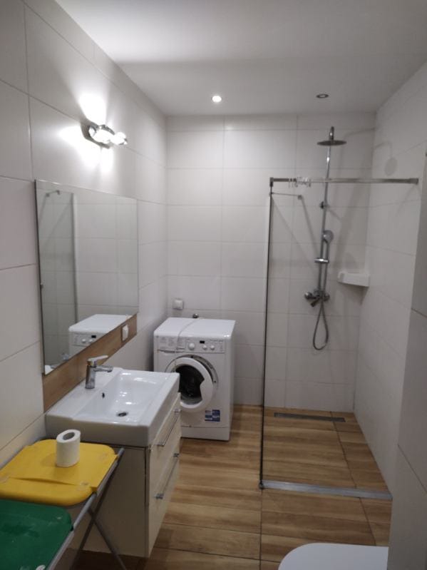 Funkcjonalna łazienka z pralką obok kabiny prysznicowej, białe płytki na ścianach, drewnopodobna podłoga i umywalka z szafką. Nowoczesne oświetlenie.