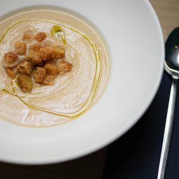 Restauracja Wola Verde - Kremowa zupa z grzankami i oliwą z oliwek w białym talerzu, obok leży srebrna łyżka na czarnej serwetce.