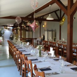 Restauracja Wola Verde - Długi stół nakryty białym obrusem z ustawionymi szklankami, sztućcami, świecami i kwiatami w wazonach, udekorowany balonami, przygotowany na uroczystość w przestronnym wnętrzu z drewnianymi...