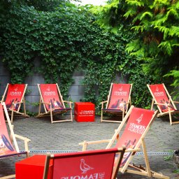 Restauracja Wola Verde - Ustawione na zewnątrz, na brukowanym patio, leżaki z drewnianą ramą i czerwonym płótnem z logo marki whisky, otoczone zielenią pnączy i drzew, tworzące strefę relaksu.