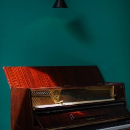 Restauracja Wola Verde - Pionowe ujęcie otwartego, brązowego pianina Legnica na tle turkusowej ściany z czarnym kinkietem, z delikatnym światłem padającym na wnętrze instrumentu.