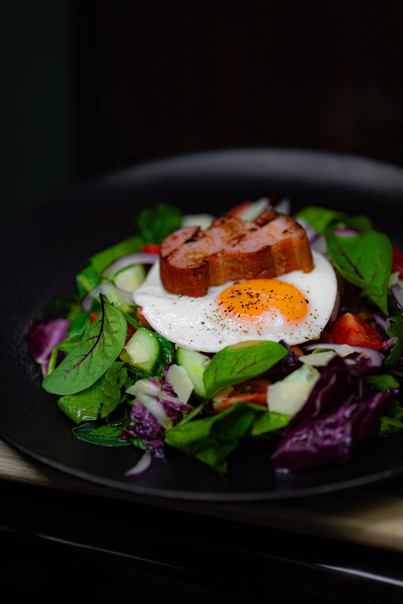 Kompozycja sałaty z jajkiem sadzonym i grillowaną kiełbasą na czarnym talerzu, z widocznymi listkami szpinaku, czerwoną cebulą i pomidorkami koktajlowymi.