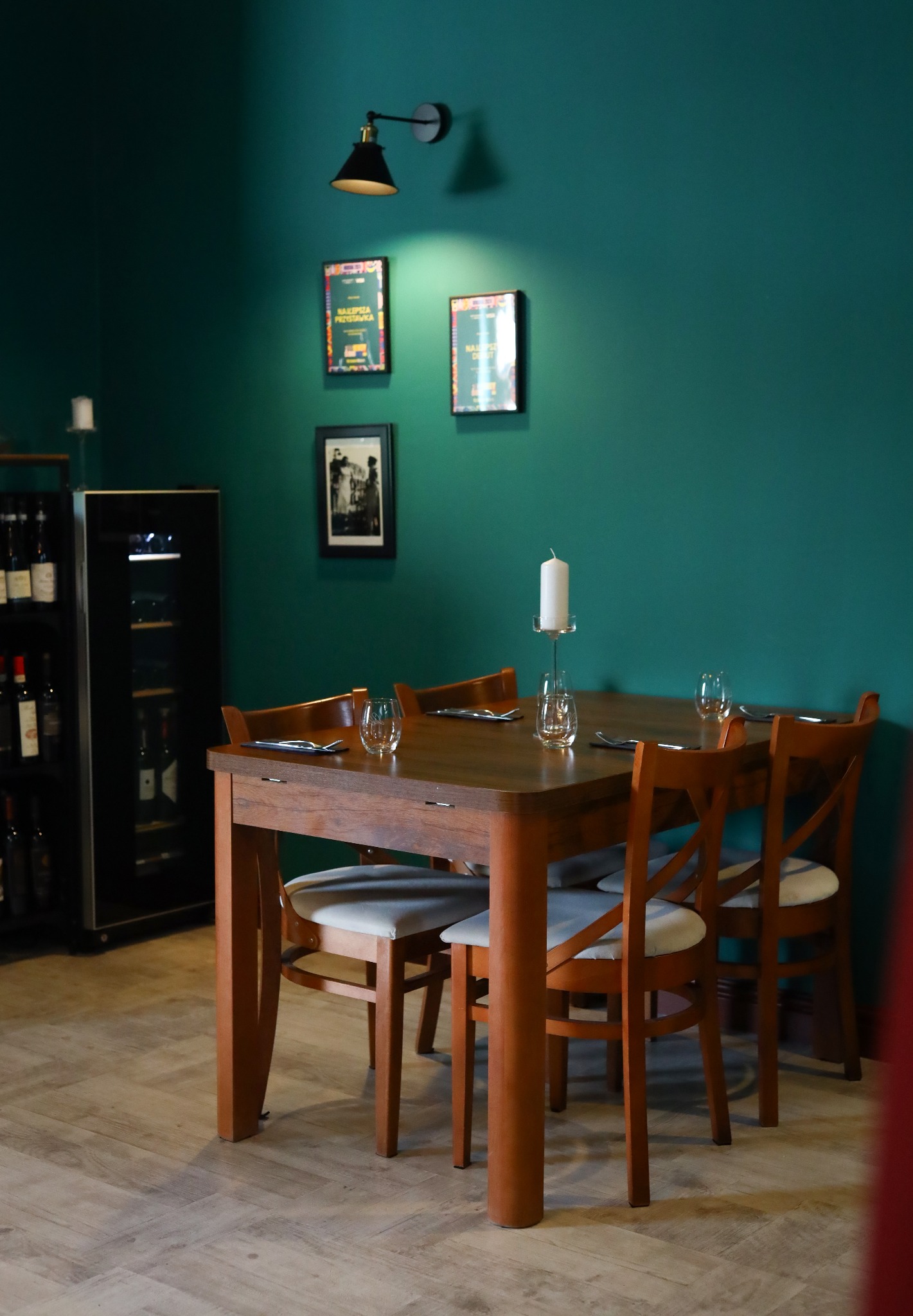 Przytulne wnętrze restauracji w Krakowie z drewnianym stołem nakrytym dla czterech osób, minimalistyczna dekoracja ze świecą i szklankami, butelki wina w tle oraz ciemnozielona ściana.