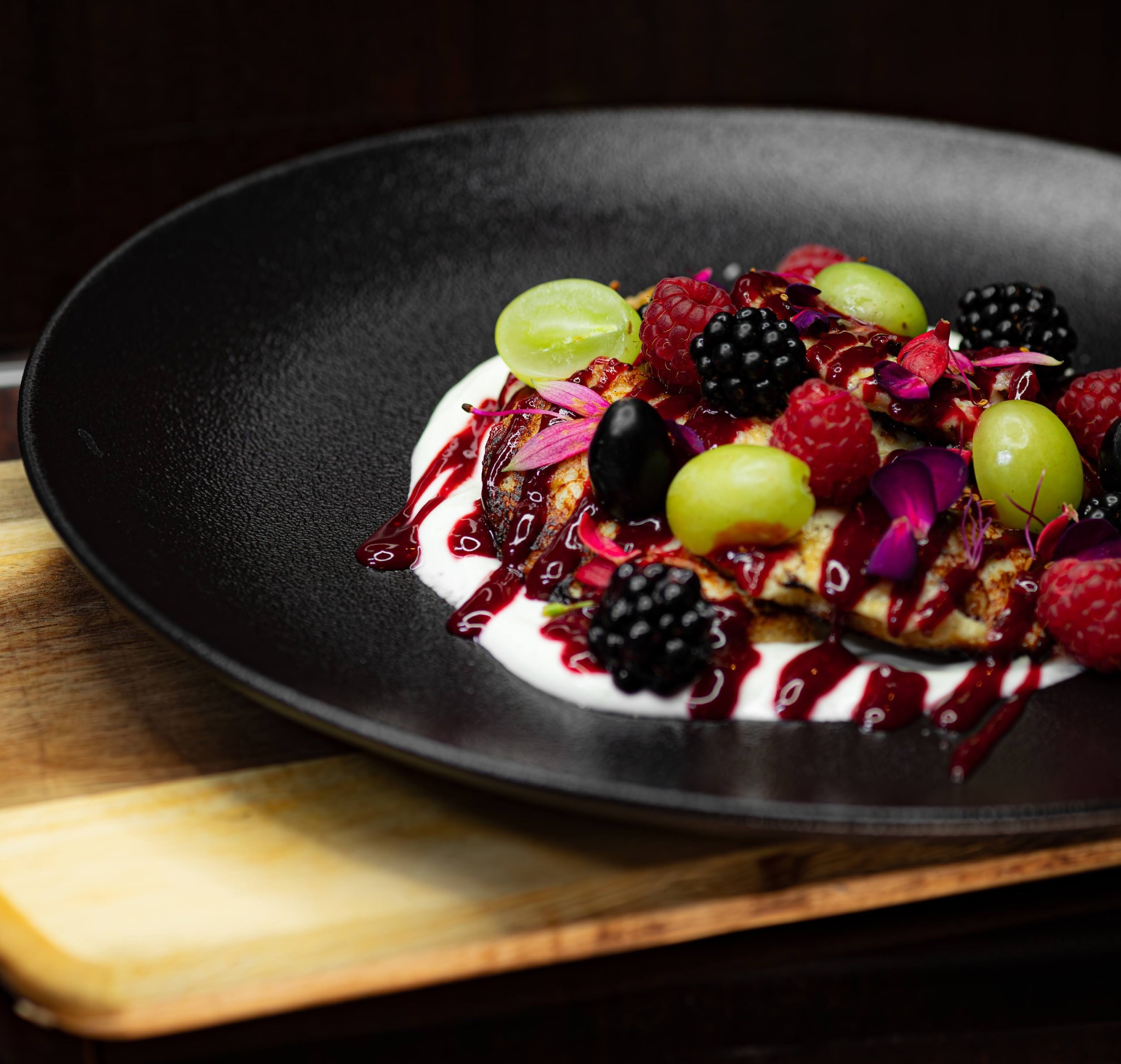 Elegancko podane naleśniki na czarnym talerzu, polane białym sosem, sosem owocowym i udekorowane świeżymi malinami, jeżynami, zielonymi winogronami oraz płatkami kwiatów.