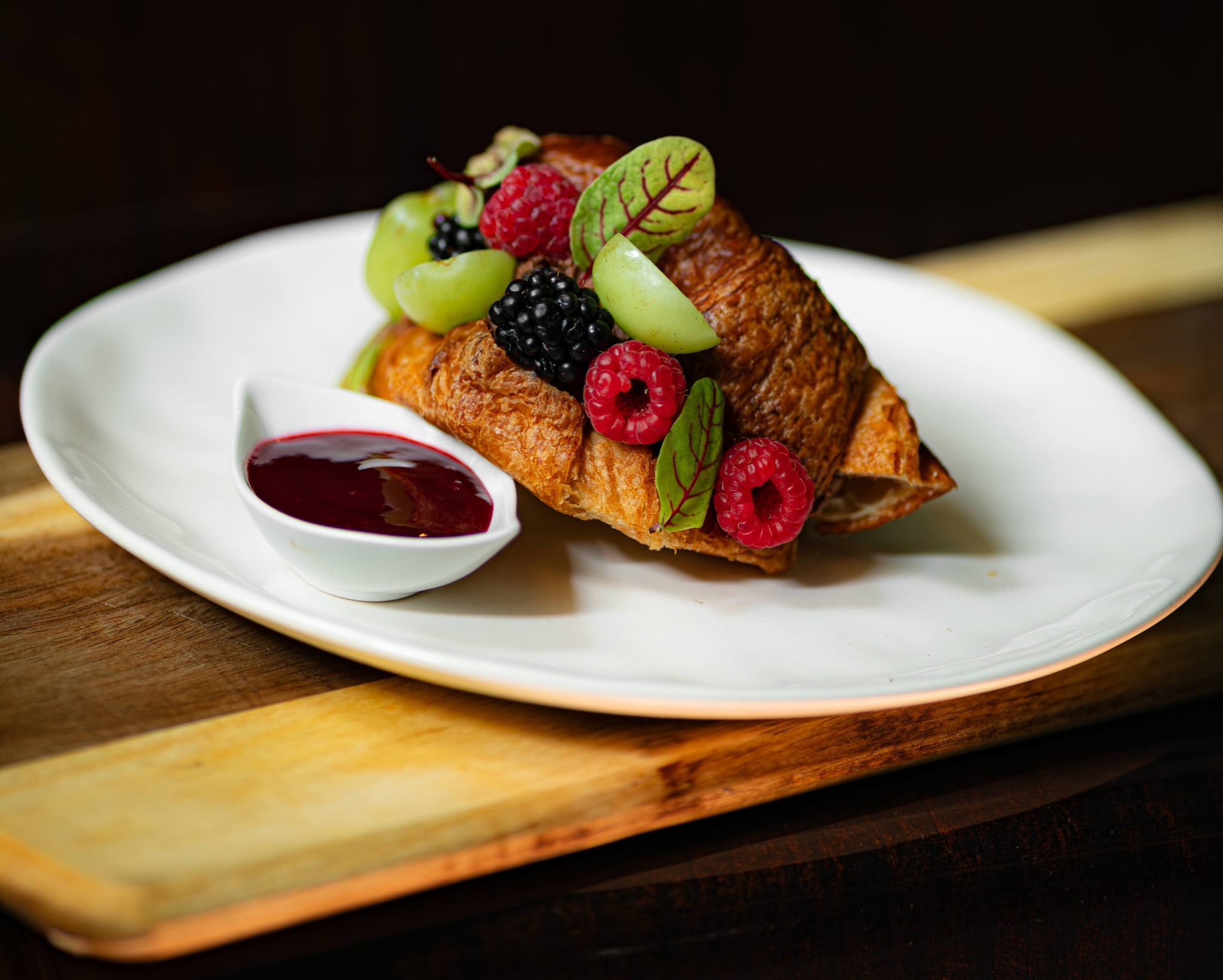Apetyczny rogalik francuski udekorowany świeżymi malinami, jeżynami, zielonymi winogronami i listkami, podany z sosem owocowym na białym talerzu, ułożonym na drewnianej desce.