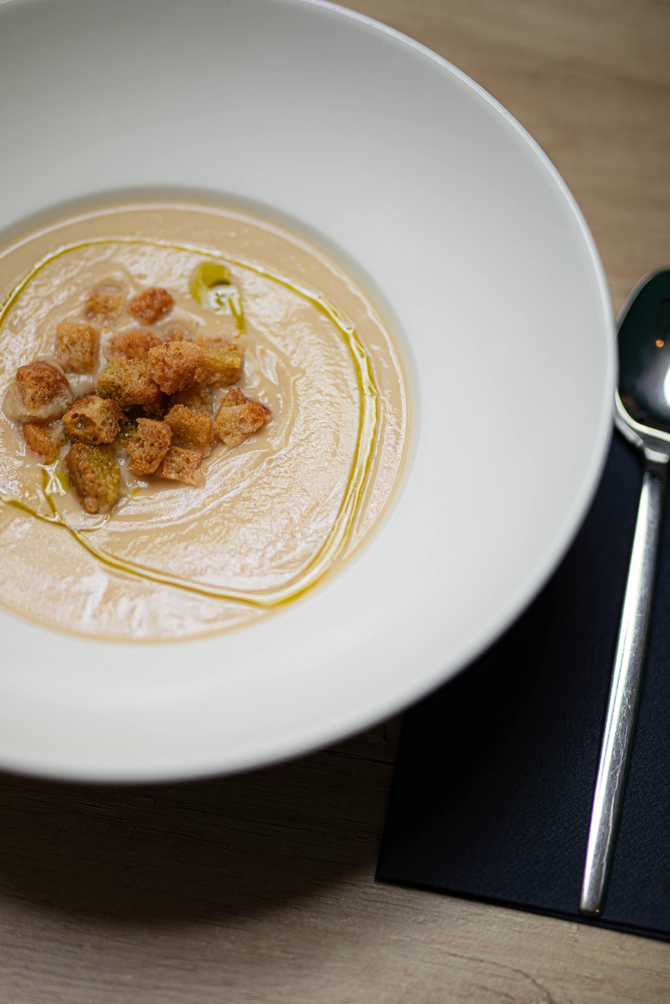 Kremowa zupa z grzankami i oliwą z oliwek w białym talerzu, obok leży srebrna łyżka na czarnej serwetce.