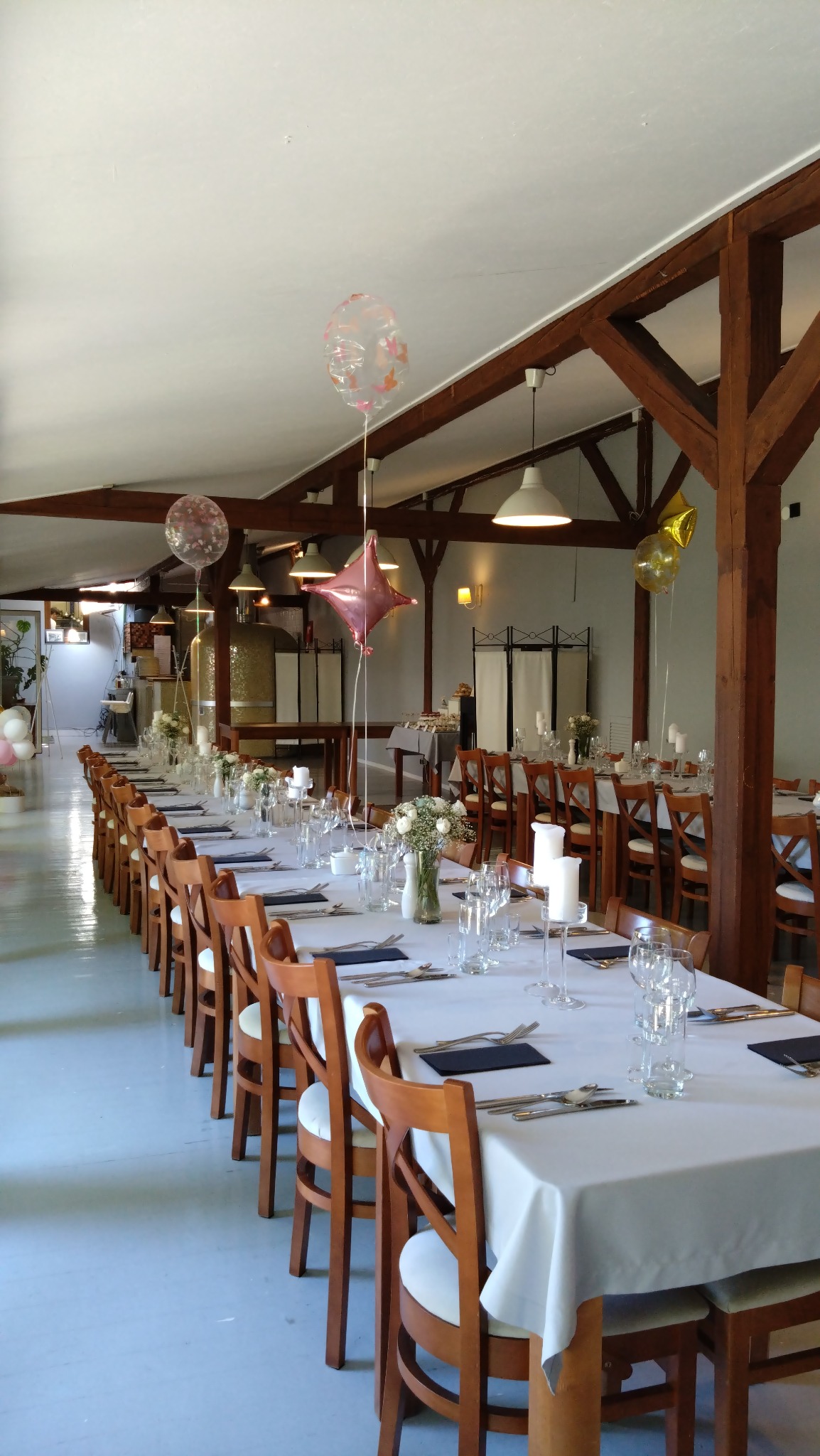 Długi stół nakryty białym obrusem z ustawionymi szklankami, sztućcami, świecami i kwiatami w wazonach, udekorowany balonami, przygotowany na uroczystość w przestronnym wnętrzu z drewnianymi...