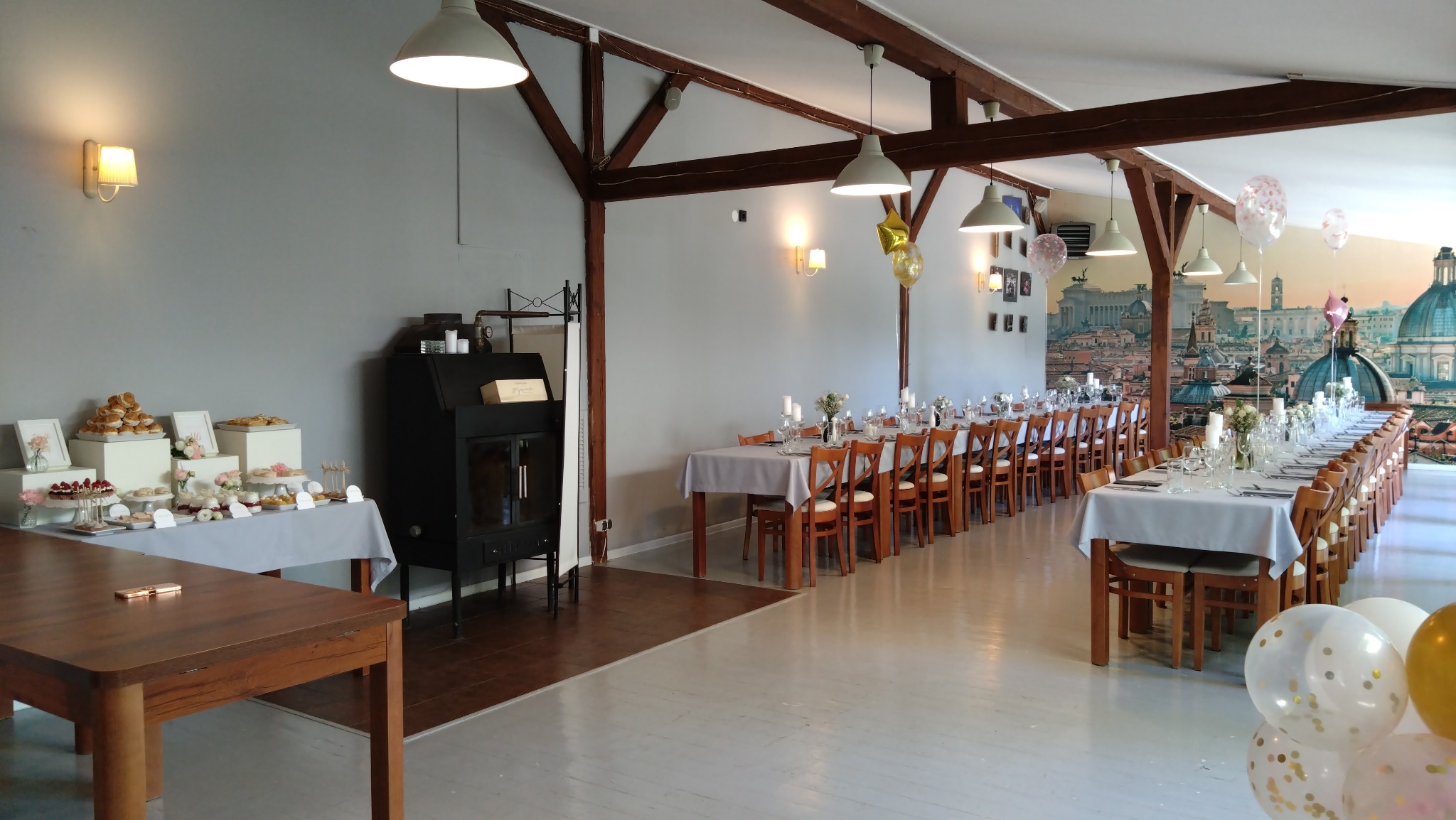 Sala bankietowa z długimi stołami nakrytymi białymi obrusami, krzesła z drewnianymi oparciami, dekoracje kwiatowe w wazonach, świece w szklanych świecznikach, balony, bufet ze słodkościami i tło...