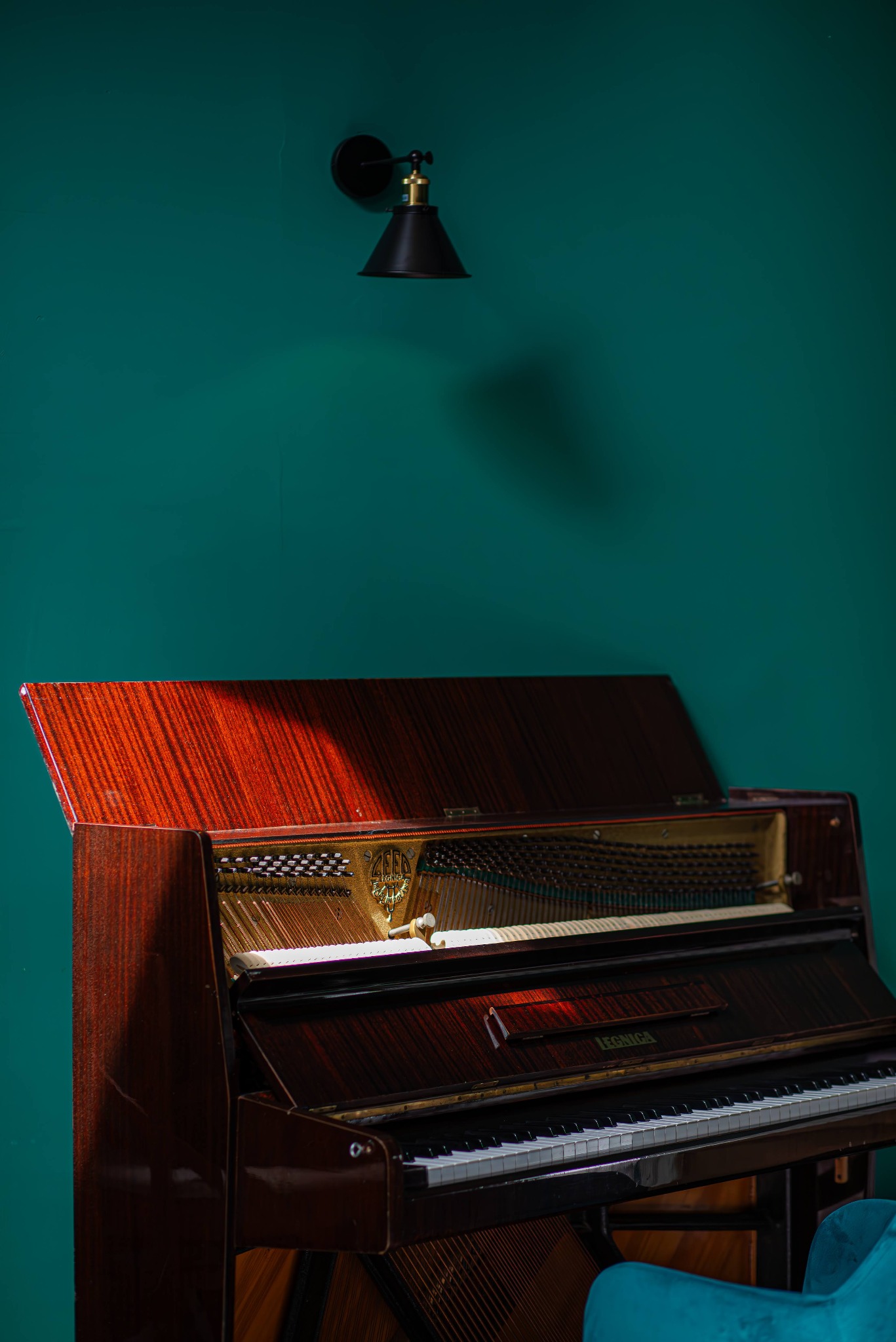 Pionowe ujęcie otwartego, brązowego pianina Legnica na tle turkusowej ściany z czarnym kinkietem, z delikatnym światłem padającym na wnętrze instrumentu.