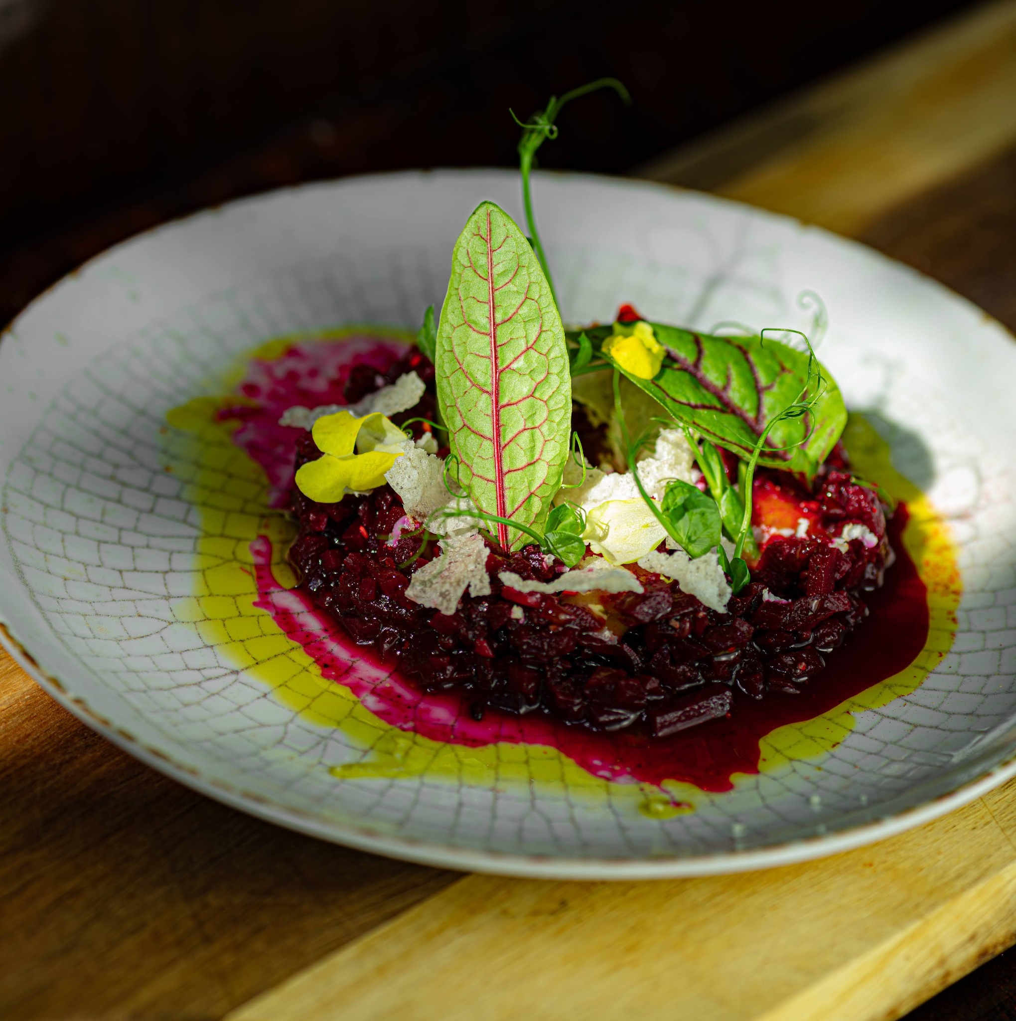 Tatar z buraka z dekoracją z liści, płatków sera i sosem na talerzu z delikatnym wzorem pęknięć, na drewnianym tle.
