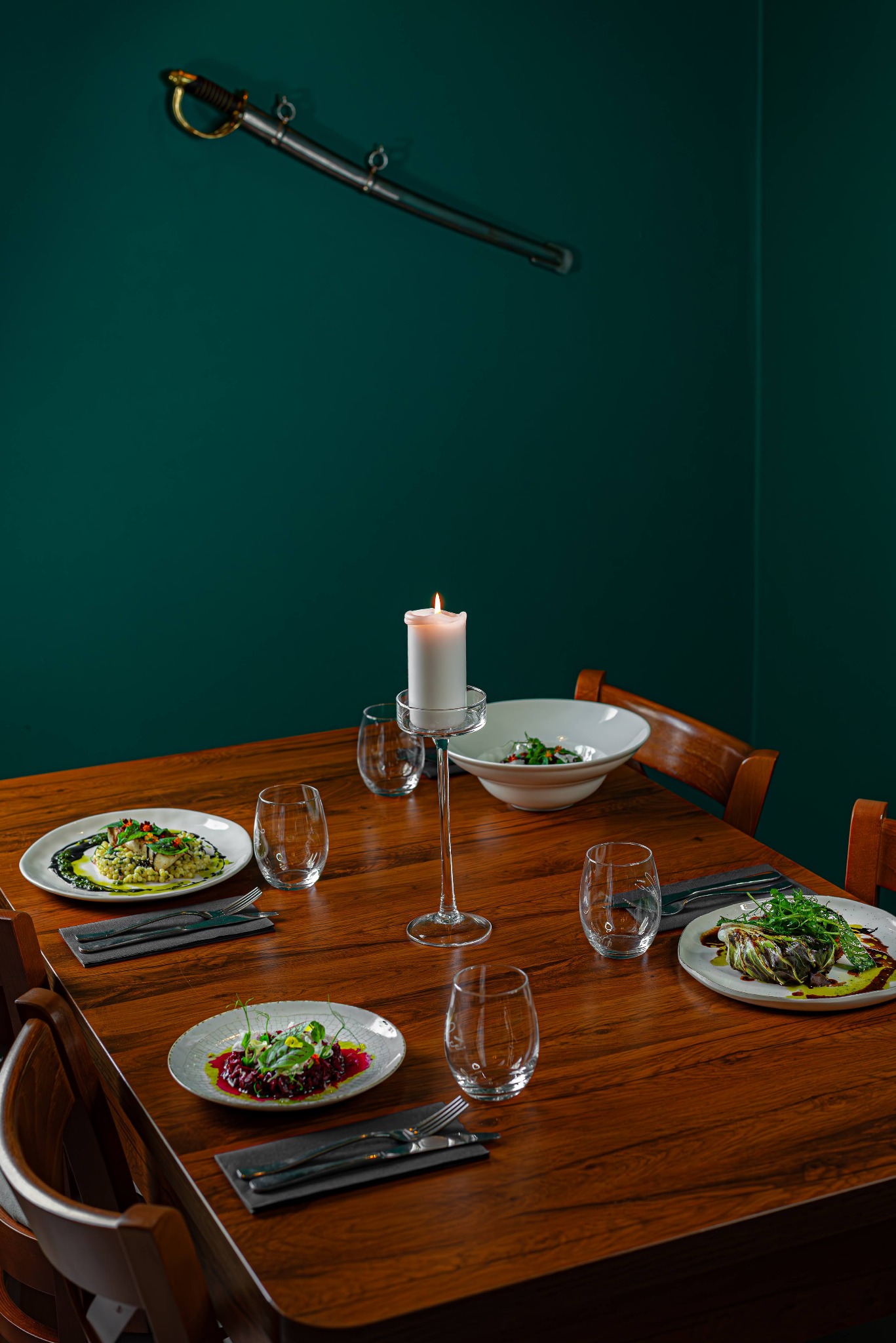 Elegancko nakryty stół z trzema talerzami z różnymi daniami, szklankami i świecą, z dekoracyjnym mieczem na zielonej ścianie w tle.